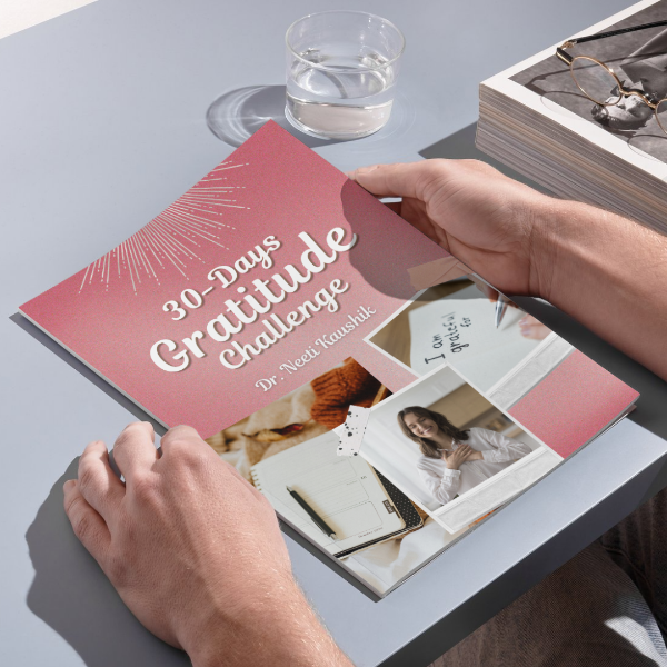 Person holding a '30 Days Gratitude Challenge' book on a table with a glass of water and photo album.